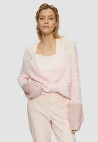 Sweter z dzianiny w odcieniach różu, z głębokim dekoltem w kształcie litery V, luźnym krojem, szerokimi rękawami oraz detalami w postaci ażurowych wzorów. Materiał wydaje się miękki i teksturowany.