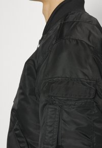 Personne portant une veste bomber noire avec une poche zippée et une poche à rabat sur la manche, le col visible sur un fond uni.