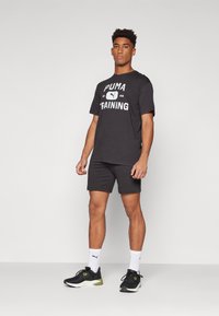 T-shirt en coton noir avec le texte "PUMA TRAINING" en blanc, associé à un short noir, des chaussettes blanches et des chaussures de sport noires avec des accents verts.