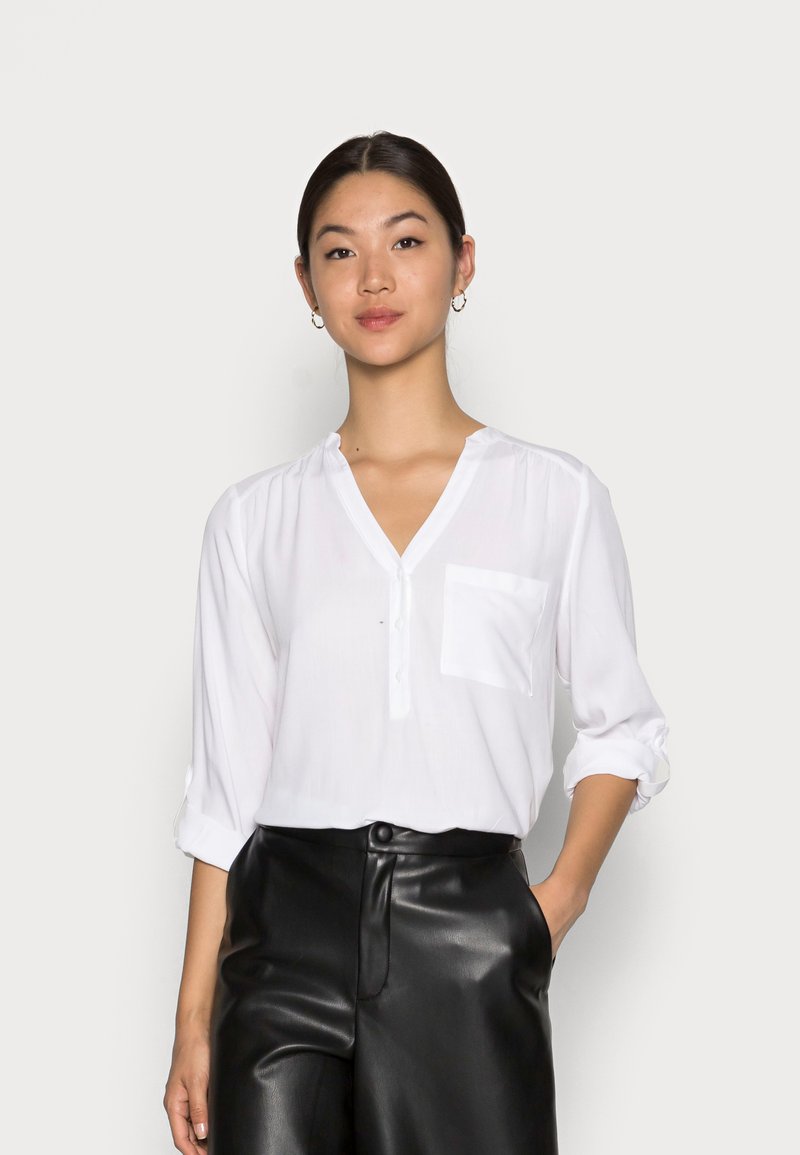 Witte blouse met V-hals, lange mouwen opgerold tot de ellebogen en een borstzak, gecombineerd met zwarte high-waisted broek.