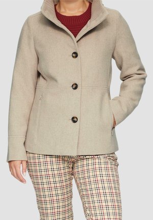 Femme portant un manteau beige boutonné, un pull rouge et un pantalon beige à carreaux, la main dans la poche, sur un fond uni.