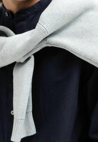 Pull en maille gris clair noué autour des épaules sur une chemise à boutons foncée, mettant en valeur un motif texturé et une couleur contrastante.
