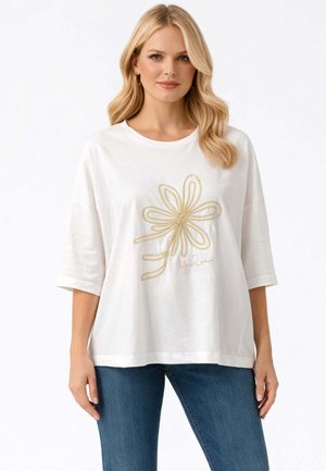 Donna bionda che indossa una maglietta bianca oversize con motivo floreale astratto beige, abbinata a jeans blu, in piedi su sfondo bianco.