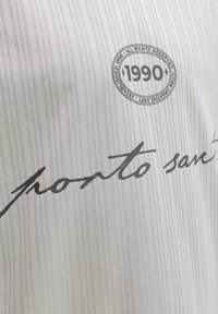Tissu blanc avec des rayures verticales légères, comportant un texte en cursive noire et un tampon circulaire indiquant "1990" avec un texte supplémentaire en petit.