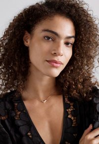 Jemný zlatý náhrdelník s přívěskem ve tvaru V. Modelka má na sobě černý texturovaný oděv s květinovými vzory a 3D akcenty. Přirozené kudrnaté vlasy.