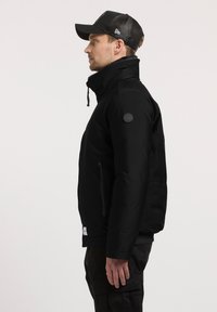 Zwarte geïsoleerde jas met hoge kraag, ritssluitingen en een gestructureerde stof; voorzien van een cirkelvormig logo op de linkerschouder.