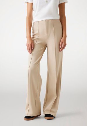 Pantaloni beige a gamba larga realizzati in tessuto morbido, con pinces frontali e vita alta. Abbinati a scarpe flat nere e marroni.