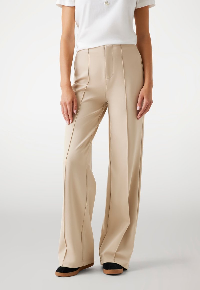 Pantalons larges beiges en tissu lisse, avec des plis avant et une taille haute. Associés à des chaussures plates noires et brunes.