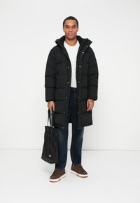 Mand med en lang sort pufferjakke, hvid t-shirt, mørke jeans, brune sneakers, som holder en sort tote-bag med logo på en hvid baggrund.