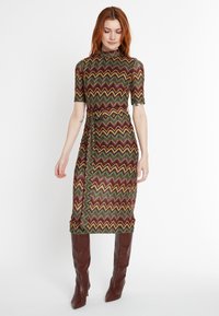 Strickkleid mit hohem Kragen und kurzen Ärmeln, das ein mehrfarbiges Zickzackmuster in Grün, Braun und Gelb aufweist. Enthält eine taillierte Linie mit Gürtel.