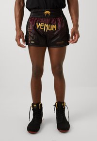 Črne in bordo atletske kratke hlače z zlatim logotipom "Venum", elastičnim pasom in rdečimi poudarnimi črtami. Nosi jih z črnimi in rumenimi čevlji.