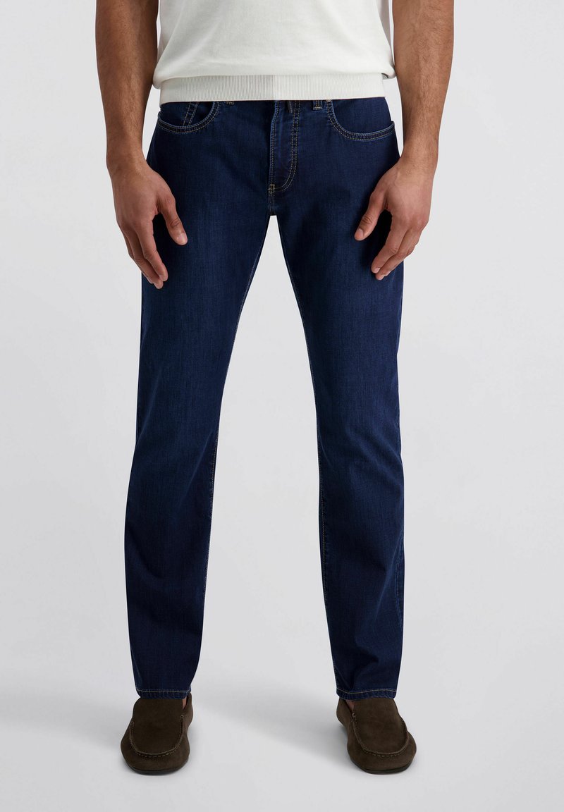 Homme portant un jean droit bleu foncé, une chemise blanche et des chaussures marron à enfiler, debout devant un fond clair uni.