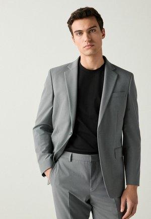 Traje gris a medida hecho de una tela suave, que presenta un blazer de un solo botón y pantalones de frente plano, combinado con una camiseta negra debajo.