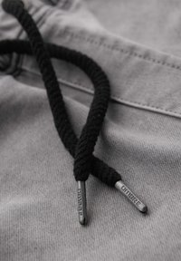 Gros plan sur les embouts noirs de lacets sur un tissu gris avec des aglets métalliques gravés du mot "OMBRE."