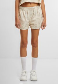 Hellbeige Shorts mit einem strukturierten geometrischen Muster, elastischem Bund und leichtem Glanz; kombiniert mit weißen knielangen Socken und Sneakers.