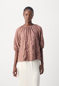 Rosa strukturierte Bluse mit aufgestellten Ärmeln und gerafftem Ausschnitt, getragen über einem fließenden weißen Rock. Glattes Material mit einem sanften Muster.
