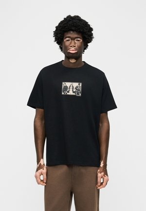 Νέος άνδρας με λεύκη φορά μαύρη γραφική μπλούζα και καφέ παντελόνια, στέκεται μπροστά σε απλό φωτεινό φόντο, κοιτάζοντας προς τα εμπρός.