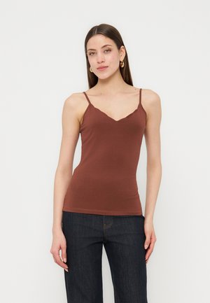 Vrouw draagt een bruin, mouwloos aansluitend topje met spaghettibandjes en donkerblauwe jeans, staand tegen een effen lichte achtergrond.