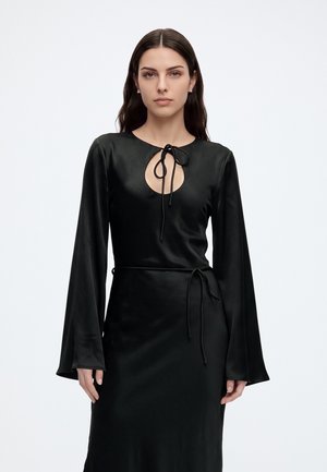Femme aux longs cheveux foncés portant une robe en satin noire avec des manches cloche, un col rond à ouverture en forme de goutte et des détails noués au niveau du cou et de la taille.