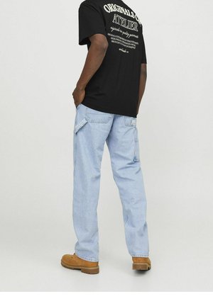 Homme portant un t-shirt noir graphique, un jean bleu clair ample avec poches latérales, et des bottes de travail marron clair, debout les mains dans les poches.