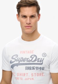 Weißes Baumwoll-T-Shirt mit Rundhalsausschnitt, mit einem grafischen Aufdruck in Grau und pinkfarbenem Text, der "Vintage SuperDry" und weitere Details zeigt.