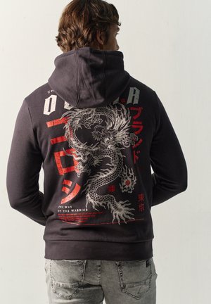 Persona con una sudadera negra con capucha con un gran diseño detallado de un dragón y caracteres japoneses rojos en la espalda, de espaldas a la cámara.