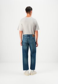 Camisa de manga corta gris claro combinada con jeans de mezclilla azul de pierna recta, que cuentan con un parche de cuero marrón y sutura sutil en el bolsillo trasero.