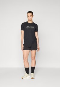 Svart treningstrøye med hvitt logo, kombinert med svarte shorts. Modellen har på seg lyse joggesko og mørke sokker. Enkel, funksjonell design.