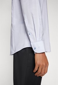 Helles blaues, langärmliges Hemd mit glatter Textur, Knopfmanschetten-Detail und abgerundetem Saum. Getragen mit dunklen Hosen.