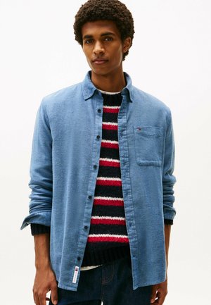 Light blue button-up shirt with a textured fabric, featuring a pocket and a small logo. Worn over a red, black, and white striped sweater.