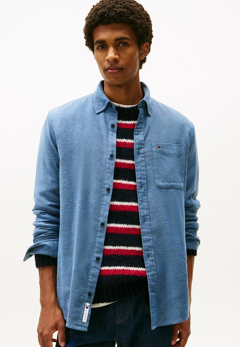 Camicia azzurro chiaro con bottoni in tessuto texturizzato, dotata di taschino e di un piccolo logo. Indossata sopra un maglione a righe rosse, nere e bianche.