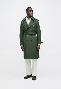 Trench coat verde con bottoni a doppio petto, ampi revers e polsini regolabili, indossato sopra pantaloni chiari e scarpe marroni.