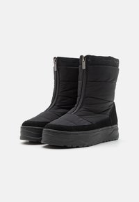 Botas de invierno acolchadas negras con cremalleras frontales, suelas gruesas y un discreto logotipo en el puño superior, mostradas una al lado de la otra sobre un fondo blanco.