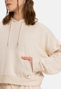 Cream-colored cropped hoodie with a drawstring hood, side pockets, and a subtle embroidered design. Soft texture and relaxed fit.