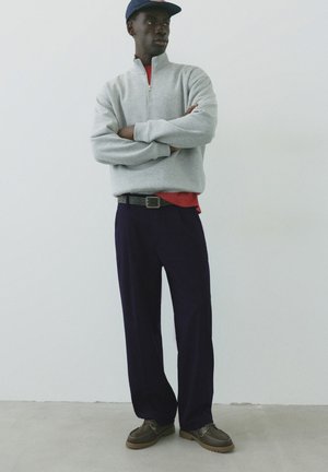 Jeune homme portant un pull gris à fermeture éclair partielle, un pantalon bleu marine, des chaussures foncées et une casquette bleue, debout les bras croisés devant un fond uni.