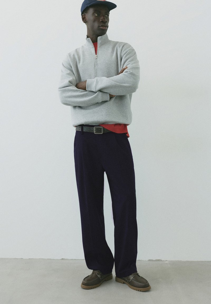 Jeune homme portant un pull gris à fermeture éclair partielle, un pantalon bleu marine, des chaussures foncées et une casquette bleue, debout les bras croisés devant un fond uni.