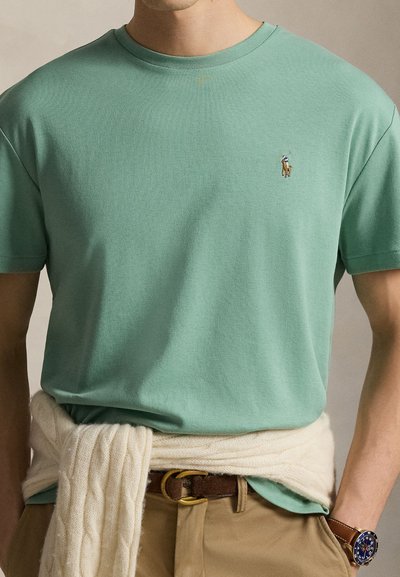 Άνδρας με ανοιχτοπράσινο μπλουζάκι με λαιμόκοψη crew και μικρό λογότυπο αλόγου, μπεζ παντελόνι με καφέ ζώνη, εκρού πλεκτή ζακέτα πλεγμένη με κόμπο στη μέση και ρολόι.