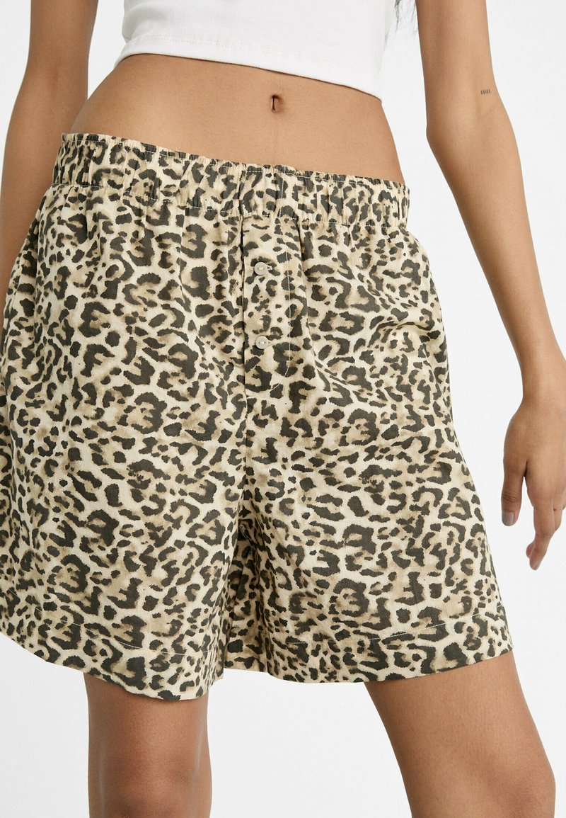 Frau in beigen und schwarzen Leopardenmuster-Shorts mit elastischem Bund und weißem Cropped-Top, stehend vor weißem Hintergrund.