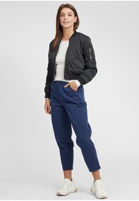 Svart bomberjacka, figursydd silhuett, ribbade muddar, ficka med dragkedja på sidan. Grå t-shirt under, marinblå byxor och vita sneakers.