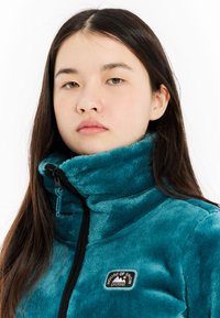 Teal fleece jack met hoge kraag en ritssluiting, voorzien van een rechthoekig logopatch. Zachte textuur, contrasterende zwarte ritsaccent.