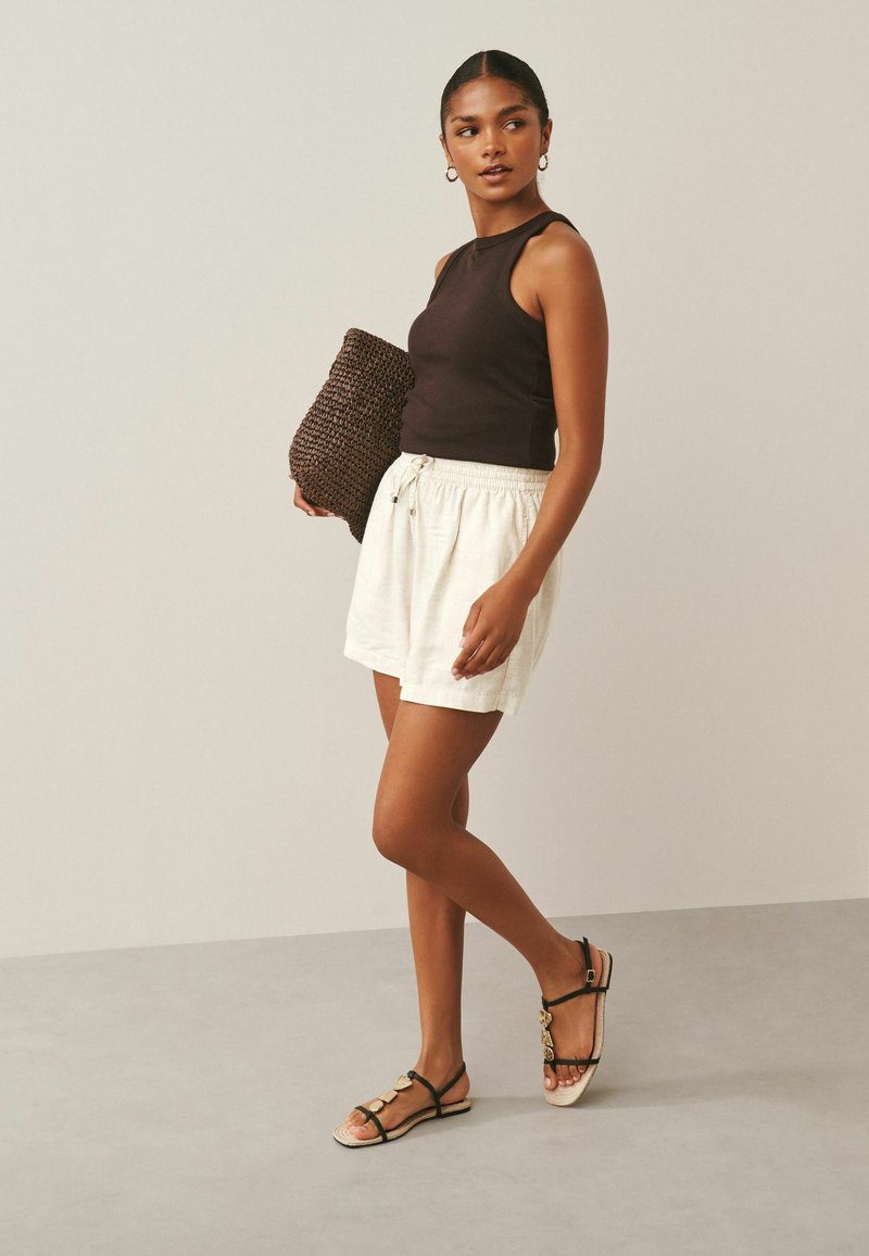 Žena v čiernom tielku bez rukávov, bielych šortkách so šnúrkou, čiernych sandáloch, drží veľkú tkanú kabelku, stojí na neutrálnom pozadí.