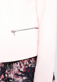 Ljusrosa texturerad jacka med ett silverfärgat dragkedjedetalj på sidan, kombinerad med en blomstermönstrad svart kjol med en samlad midja.