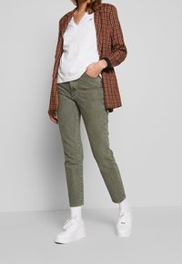 Kvinna som bär olivgröna jeans, vita sneakers och strumpor, vit t-shirt med v-ringning och brun rutig kavaj, står med en hand i fickan.