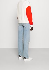 Felpa bianca con maniche arancioni fluorescenti, abbinata a jeans a gamba larga blu chiaro e sneakers bianche con dettagli neri.