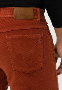 Pantaloni in corduroy color ruggine con tasca posteriore e patch in pelle marrone, indossati da una persona in maglietta nera.