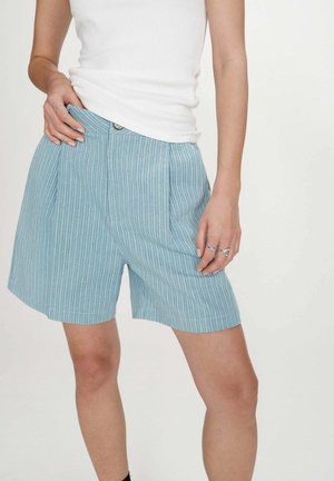 Femme portant un short rayé bleu et blanc et un haut sans manches blanc, montrant la position des mains et des bagues argentées aux doigts.