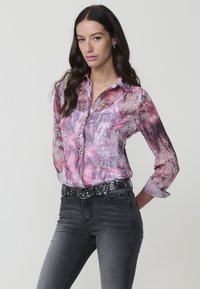 Camisa con un estampado abstracto colorido en tonos de rosa y morado; de manga larga, con cuello y tela semitranslúcida; combinada con jeans gris oscuro.