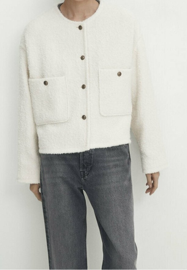 Biała, puszysta, krótka kurtka z okrągłym dekoltem, dwoma dużymi kieszeniami z przodu i pięcioma brązowymi guzikami na środku. Noszona na ciemnych dżinsach.