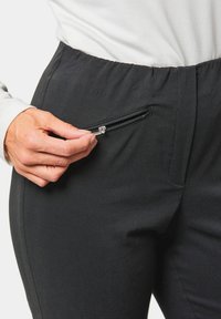 Zwarte broek met een gladde textuur, voorzien van een ritszak aan de voorkant en een rekbare tailleband. Close-up van een hand die naar de rits reikt.