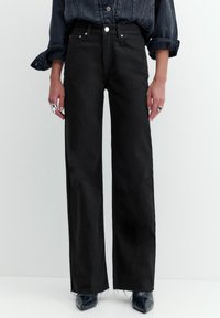 Osoba nosząca czarne dżinsy z wysokim stanem i szerokimi nogawkami, z surowymi wykończeniami, ciemną denimową koszulę z podwiniętymi rękawami oraz czarne, spiczaste buty.